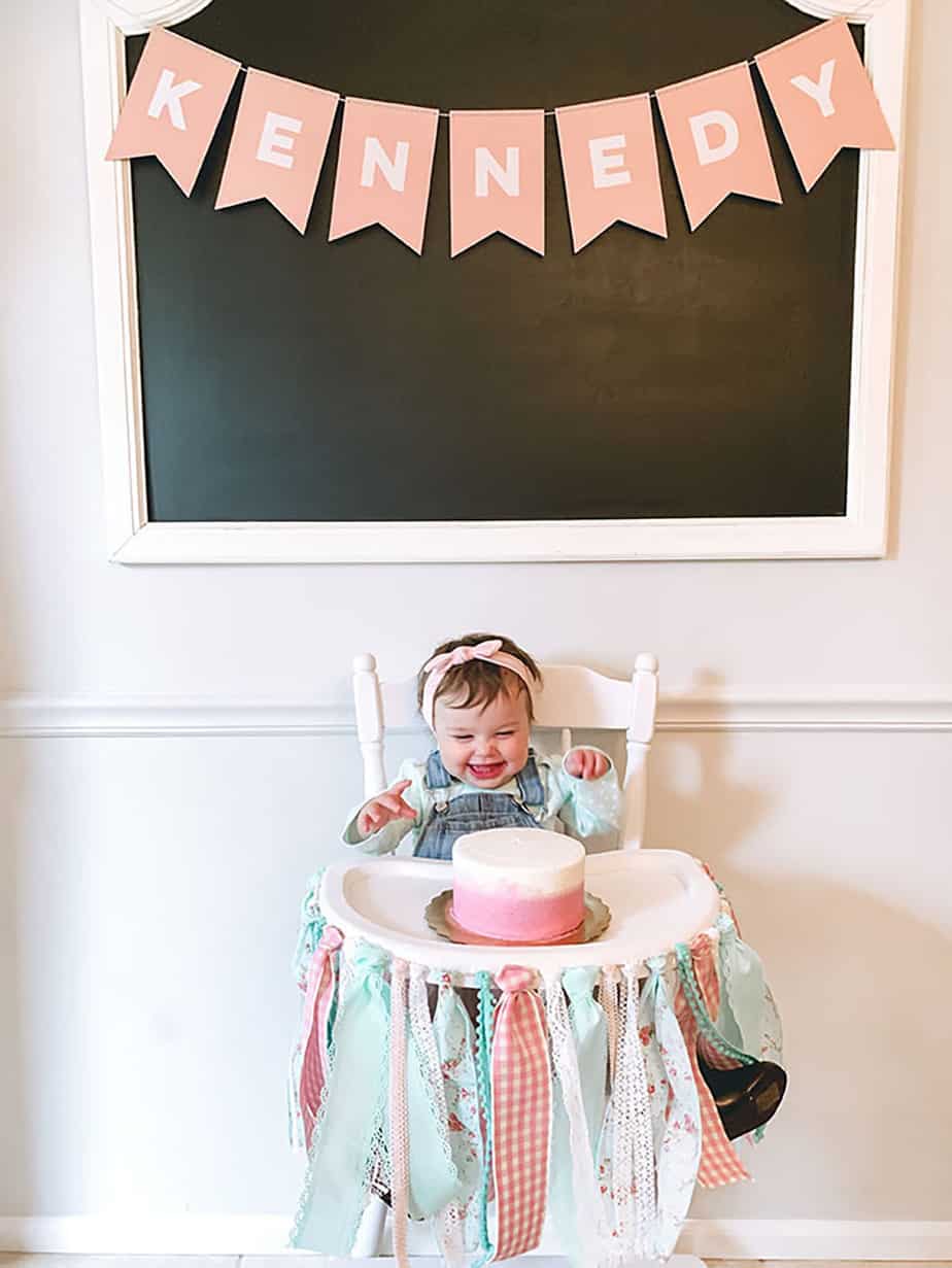 pink name banner says "kennedy" with a white highchair in front and little girl trying cake