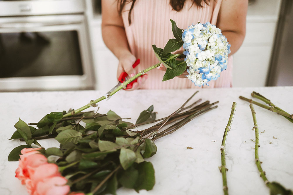 Woman wearing pink top holding floral scissors cutting blue hydrangeas and pink roses