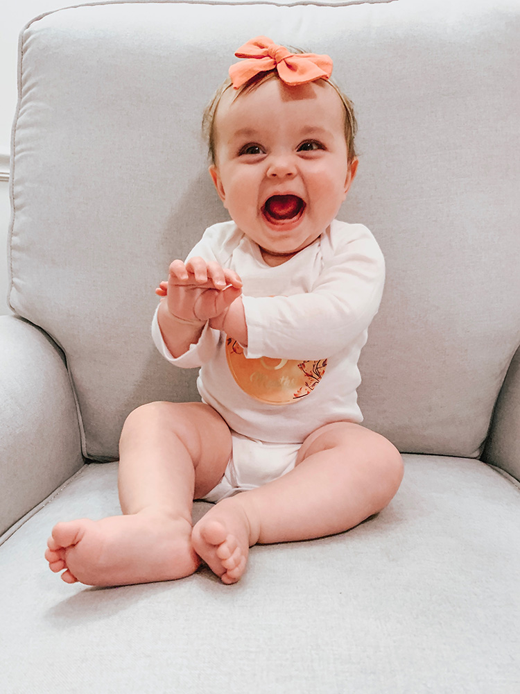 little girl sitting in grey chair with 8 month sticker on her white onesie