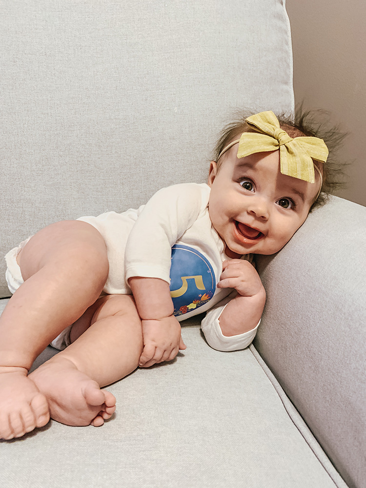little girl sitting in grey chair with 5 month sticker on her white onesie