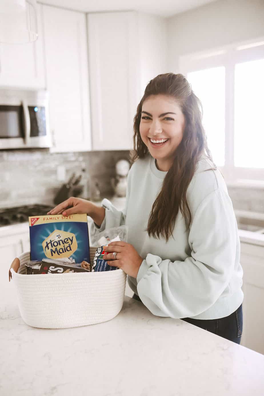 woman wearing blue sweater standing in white kitchen holding a white rope basket full of smores ingredients