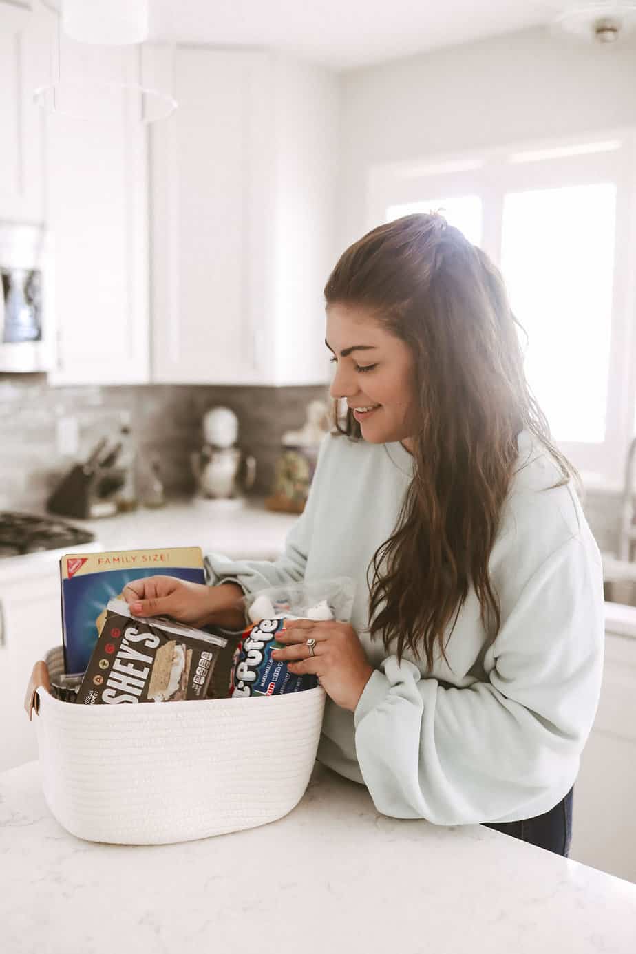 woman wearing blue sweater standing in white kitchen holding a white rope basket full of smores ingredients