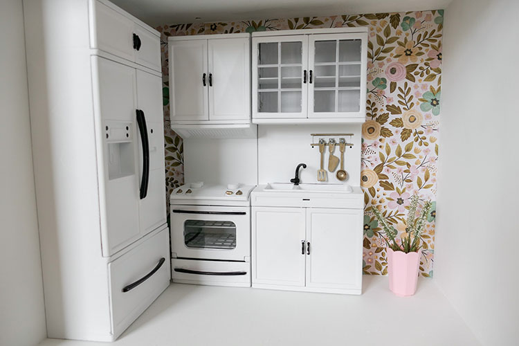 DIY dollhouse makeover project using a thrifted bookshelf showing black and white kitchen cabinets and floral wallpaper.