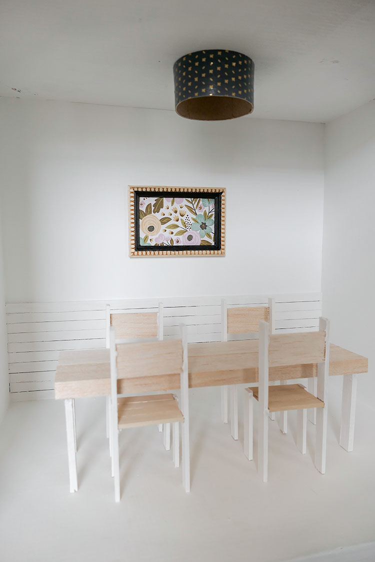 DIY dollhouse makeover project using a thrifted bookshelf showing dining room with white shiplap wall, wood dining table, framed floral artwork