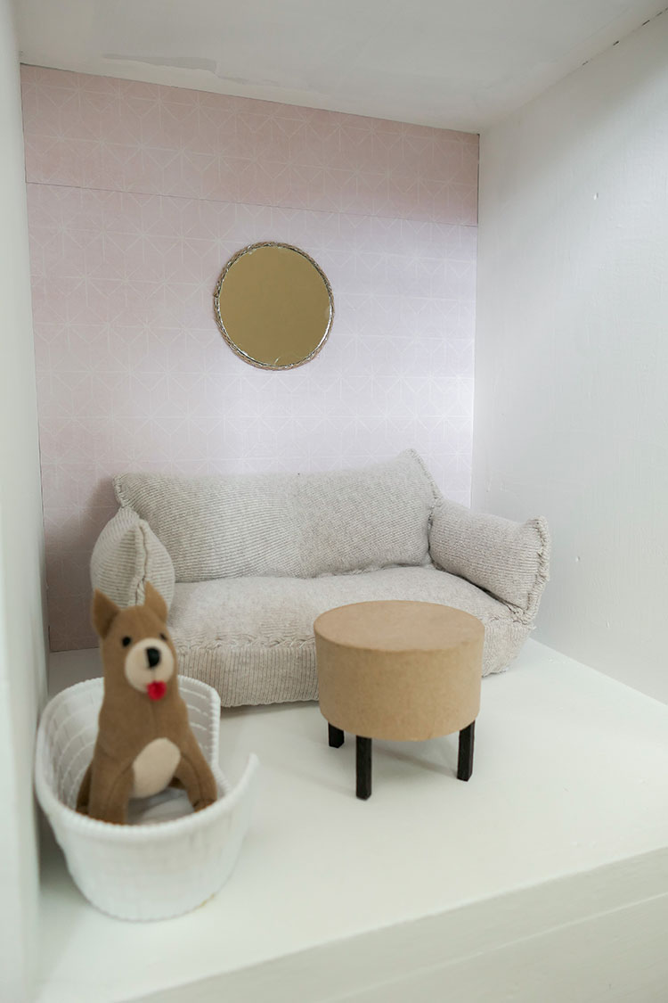 DIY dollhouse makeover project using a thrifted bookshelf showing living room with pink wallpaper and cream couch.