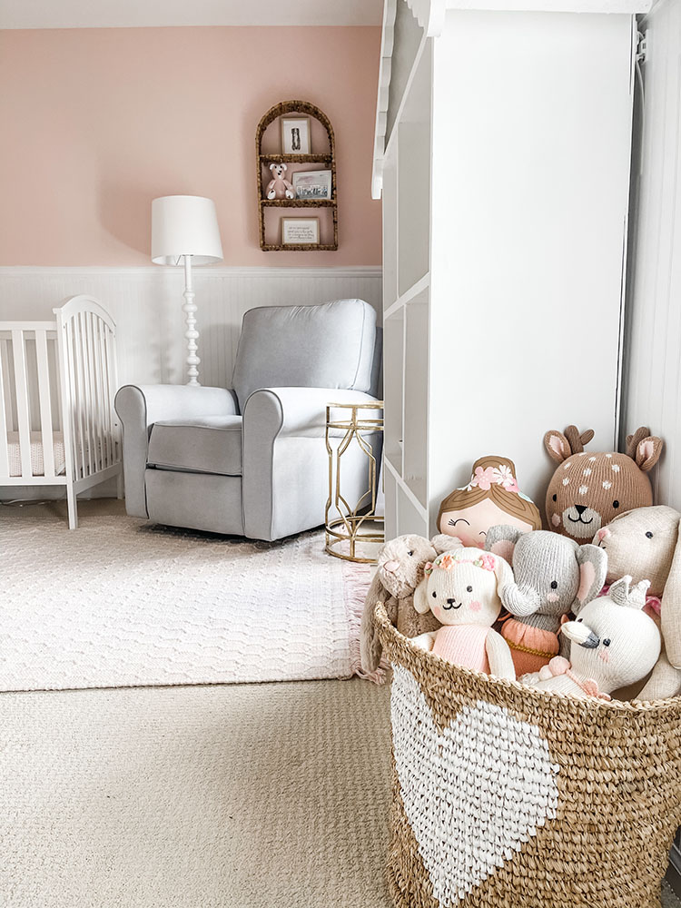 Wicker basket with white heart on it full of little girl's stuffed animals and dolls with grey pottery barn glider in the background