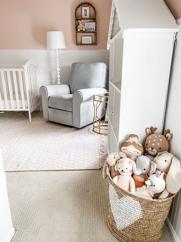 Wicker basket with white heart on it full of little girl's stuffed animals and dolls with grey pottery barn glider in the background