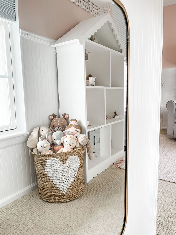 Wicker basket with white heart on it full of little girl's stuffed animals and dolls reflecting in a gold mirror
