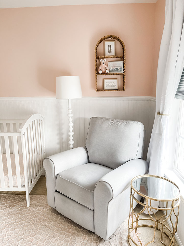 Little girl's pink and white bedroom showing grey pottery barn glider, gold side table, wicker hanging shelf, white lamp
