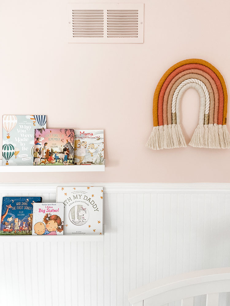 Little girl's pink and white bedroom with white shelves on the wall for books and macrame rainbow art