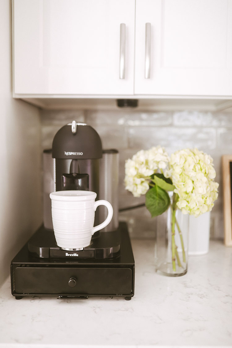 Nespresso coffee maker on white countertop filling up a white coffee cup next to vase of white hydrangeas