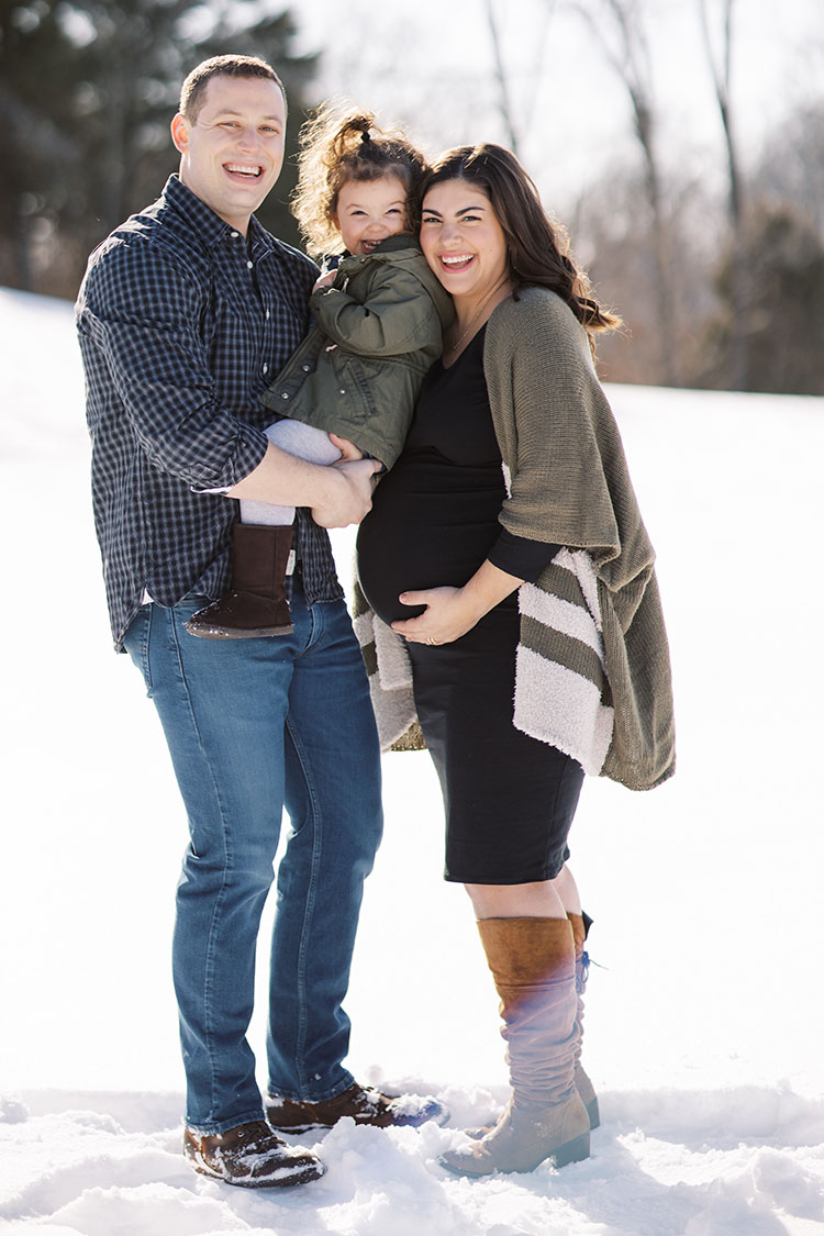 Dad, pregnant mom, and toddler girl smiling in the snow