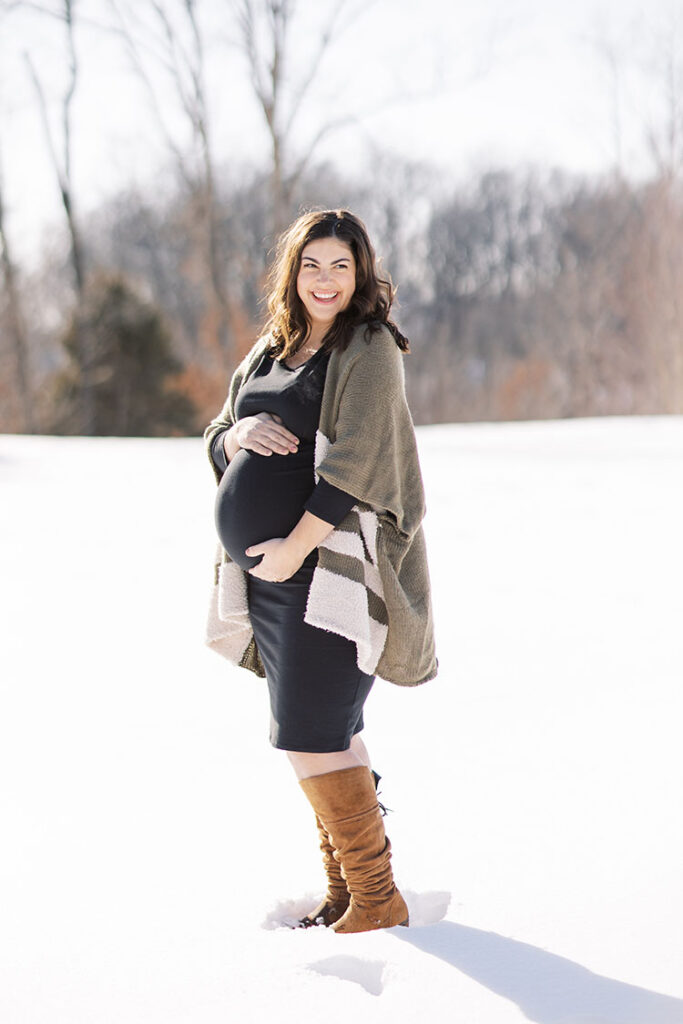 Pregnant mom wearing black dress, tall boots, and green and white striped sweater standing in the snow