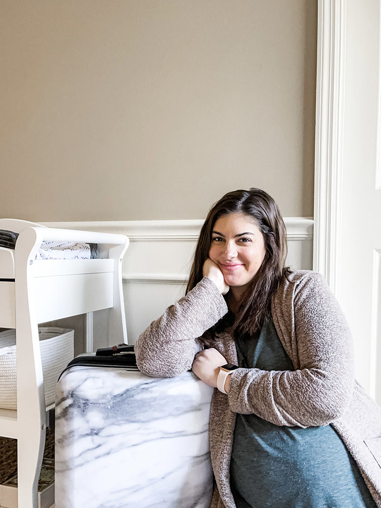 Pregnant mom in green shirt and brown sweater leaning on white marbled suitcase