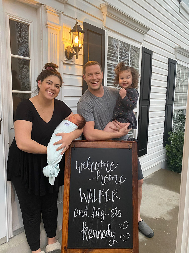 Family of four posing in front of chalkboard welcoming home a new baby to the family.