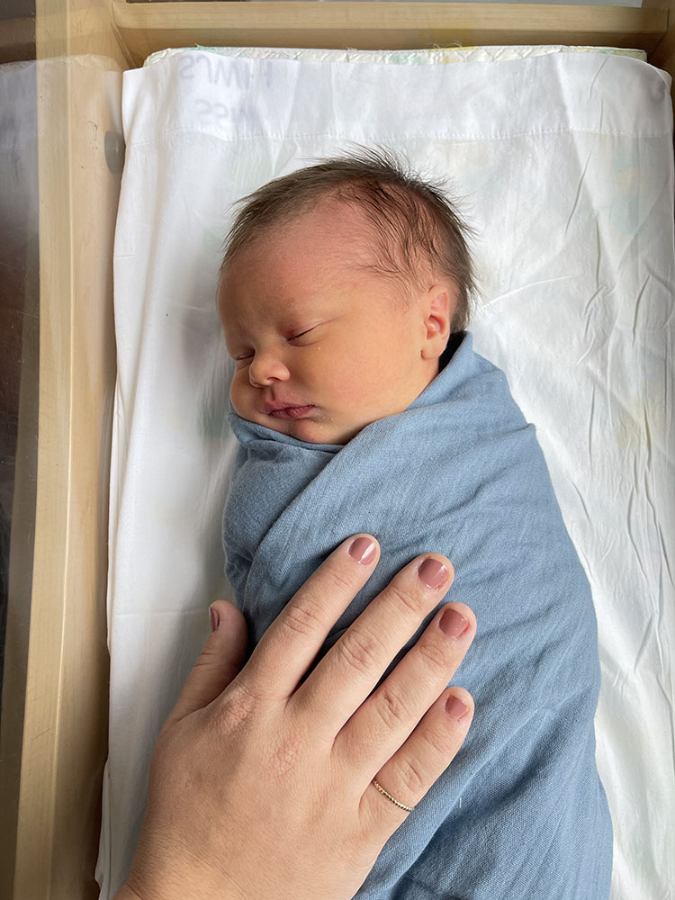 Newborn baby boy swaddled in blue blanket laying in hospital bassinet with mom's hand on his chest.