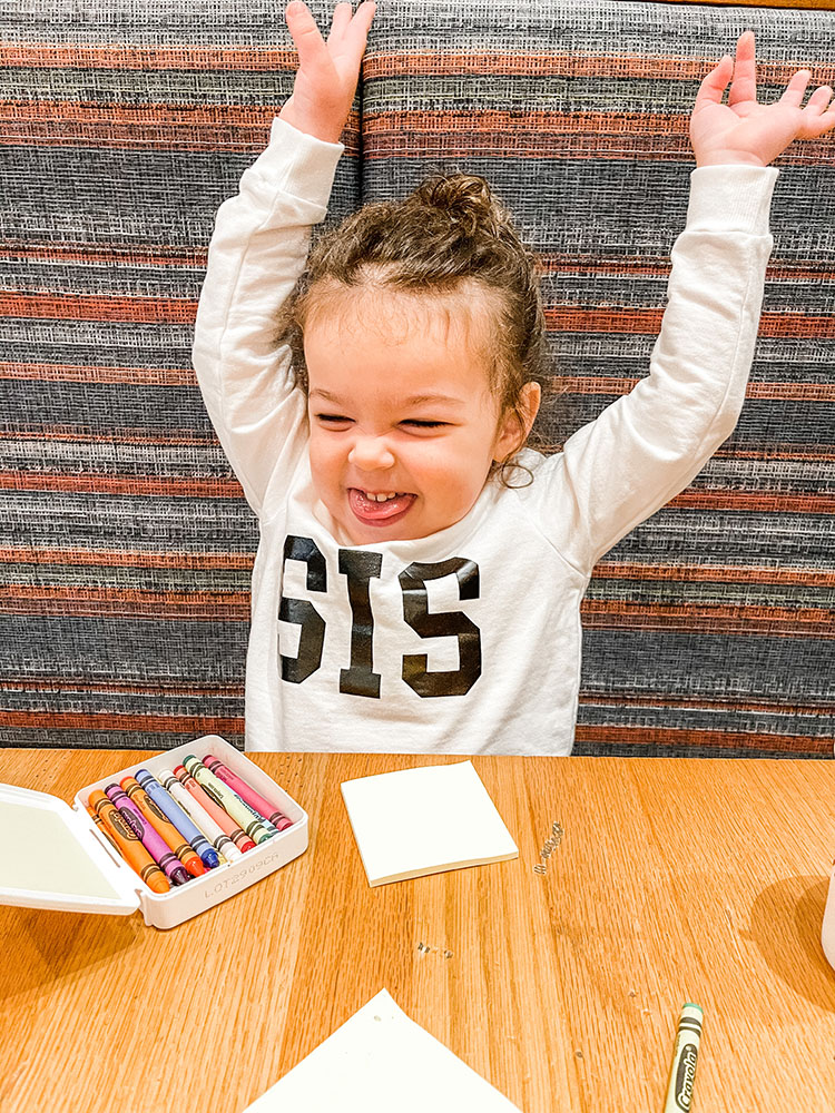 Little girl wearing "sis" sweatshirt coloring at a table.