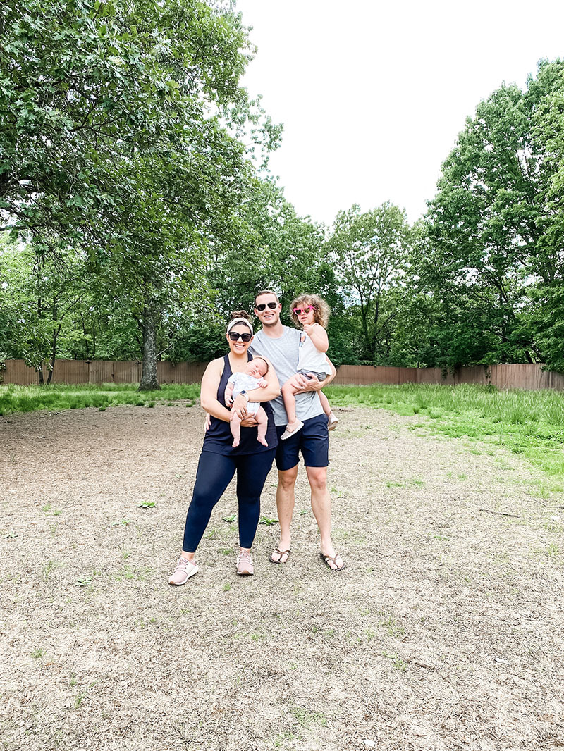 Family of four standing in the middle of a yard where a new home build will take place surrounded by trees.