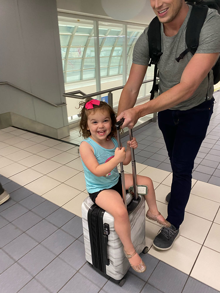Little girl riding on suitcase being pushed by dad through an airport.