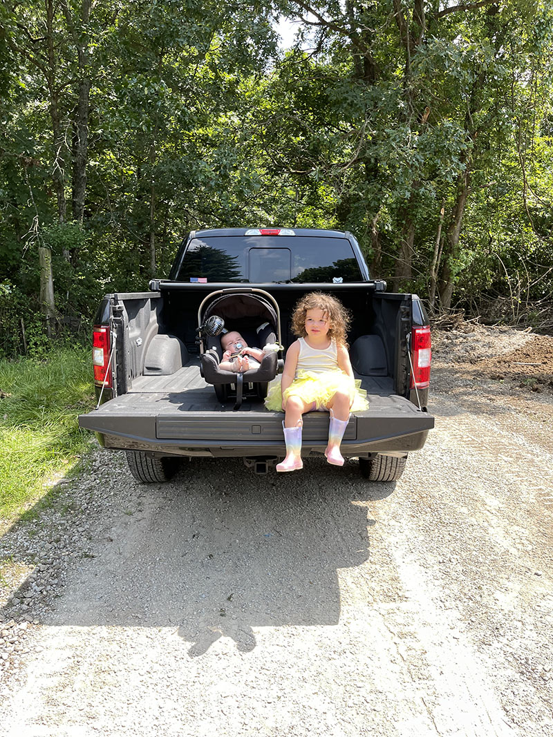 Little girl and baby sitting in back of a Ford truck bed surrounded by trees on a gravel road.