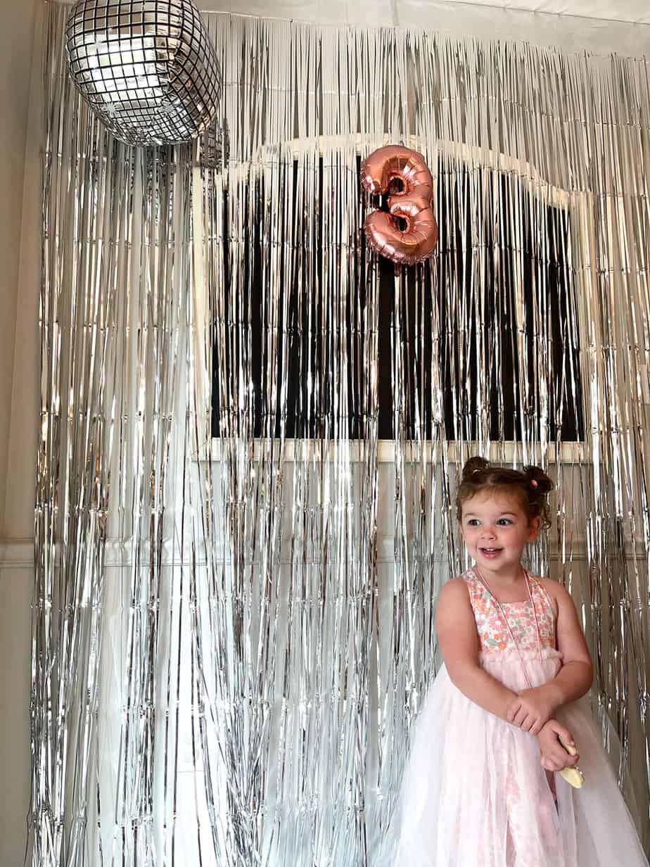 Metallic fringe photo backdrop with disco ball balloon and rose gold "3" balloon celebrating a 70s themed third birthday party.