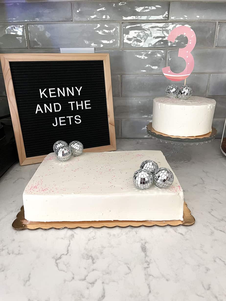 White sheet cake with sprinkles and disco balls next to smaller white cake on a glass cake stand with #3 cake topper.