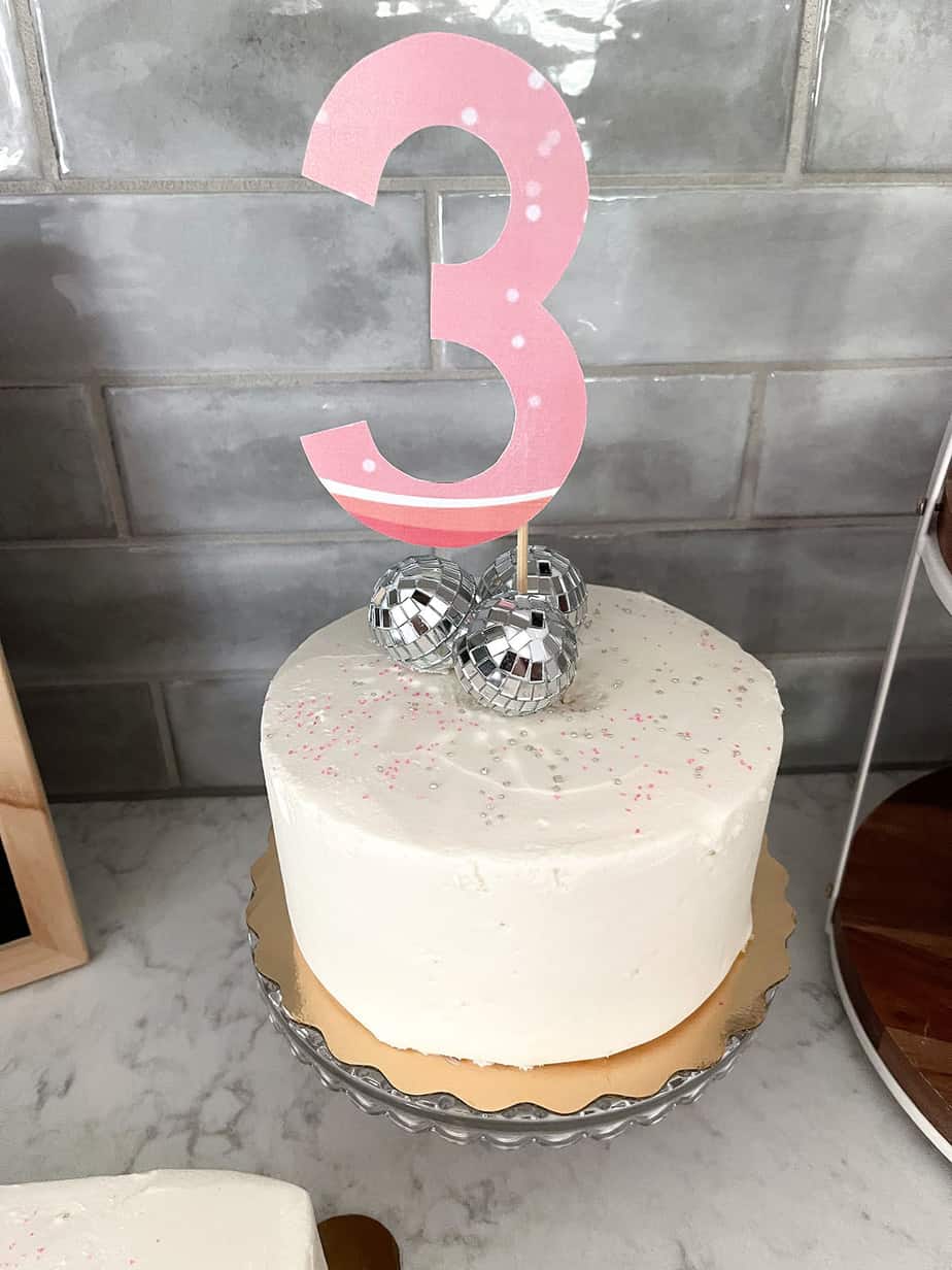 Small white cake with pink and silver sprinkles, disco balls, and a large #3 standing upright on a wooden skewer.