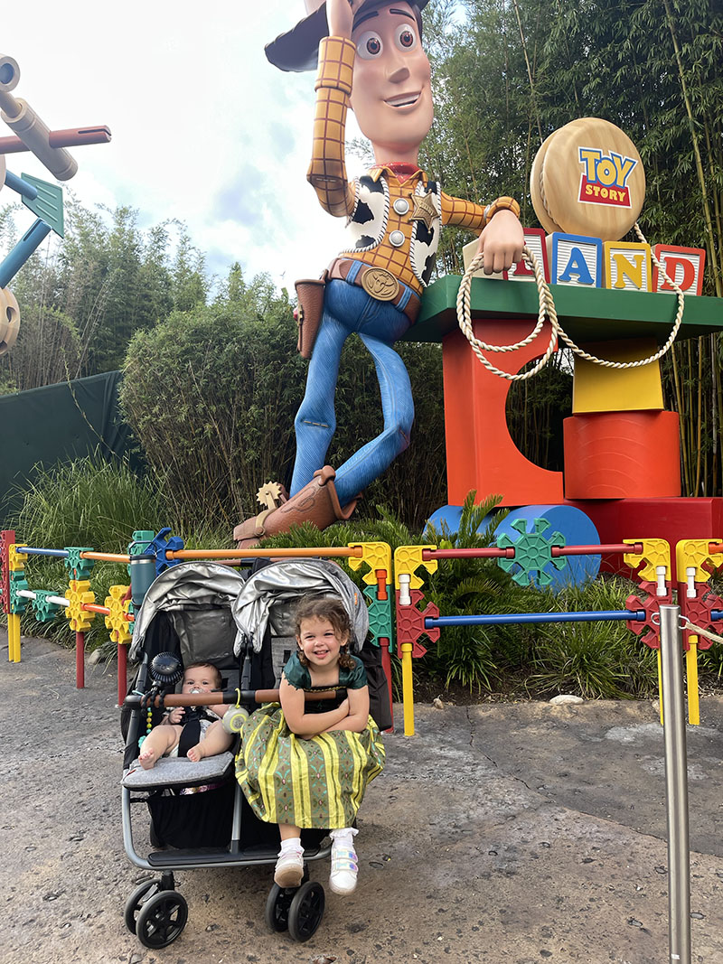 Little girl in Princess Anna dress and baby boy sitting in Zoe Twin+ Double Stroller posing in front of Toy Story Land at Disney World.