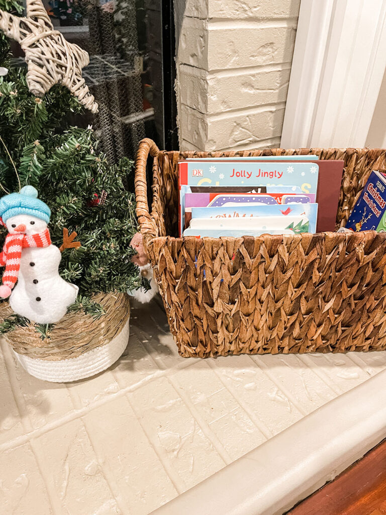 Family holiday book basket on the ledge of fireplace with holiday books inside next to a mini Christmas tree with snowman ornament.