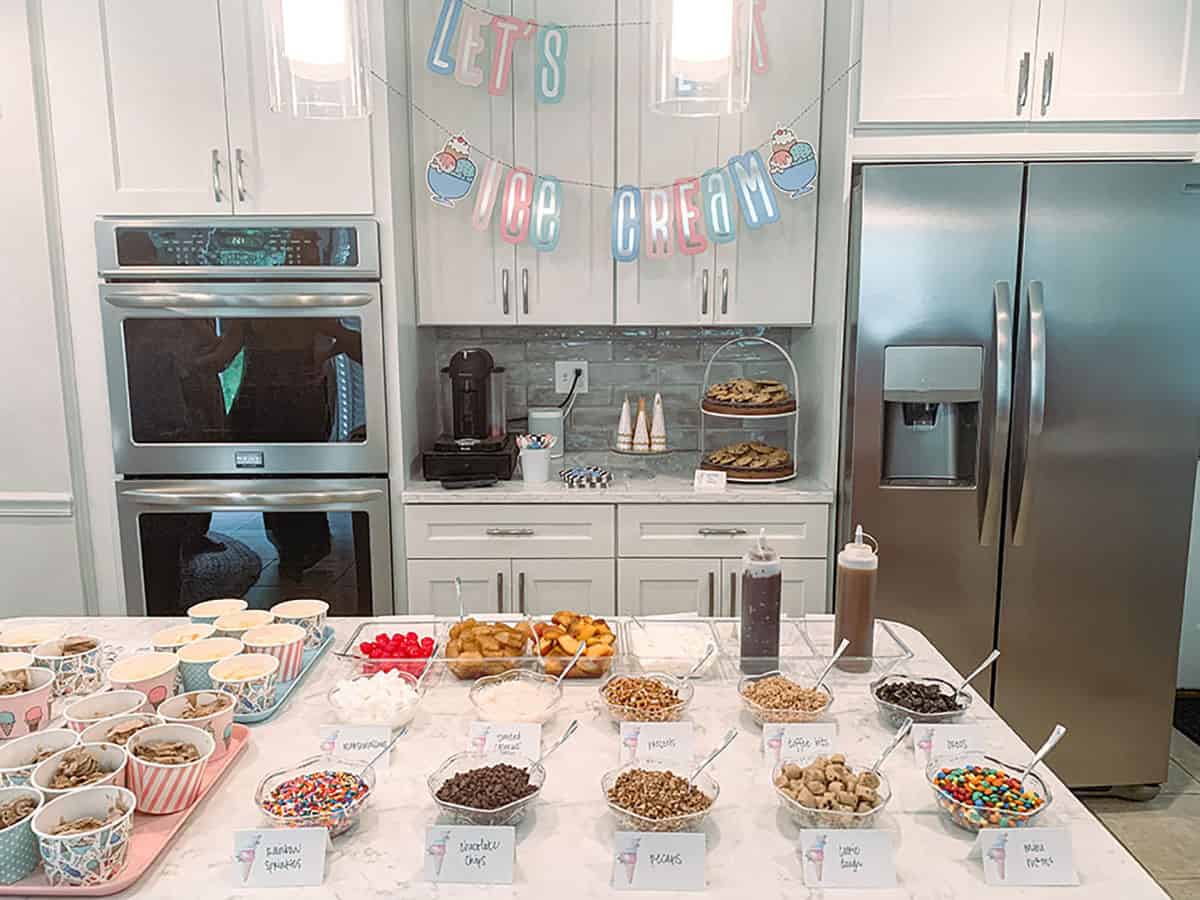 Kitchen set up for ice cream social with mini bowls of ice cream toppings (sprinkles, cookie dough, candies, pretzels, cookies) along with fresh fruit and syrup.