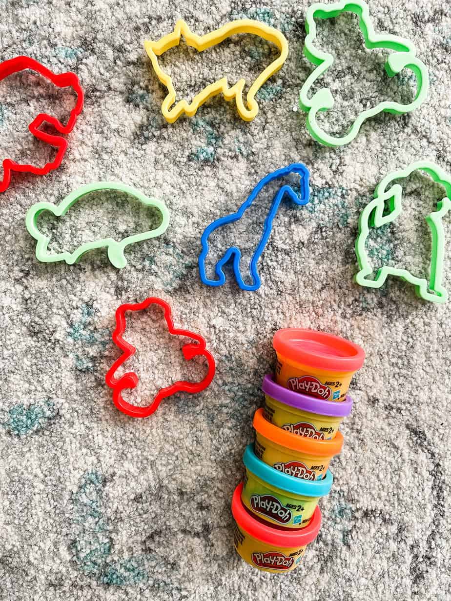 Colorful animal shaped cookie cutters and mini tubs of playdoh laid out next to one another on top of rug.