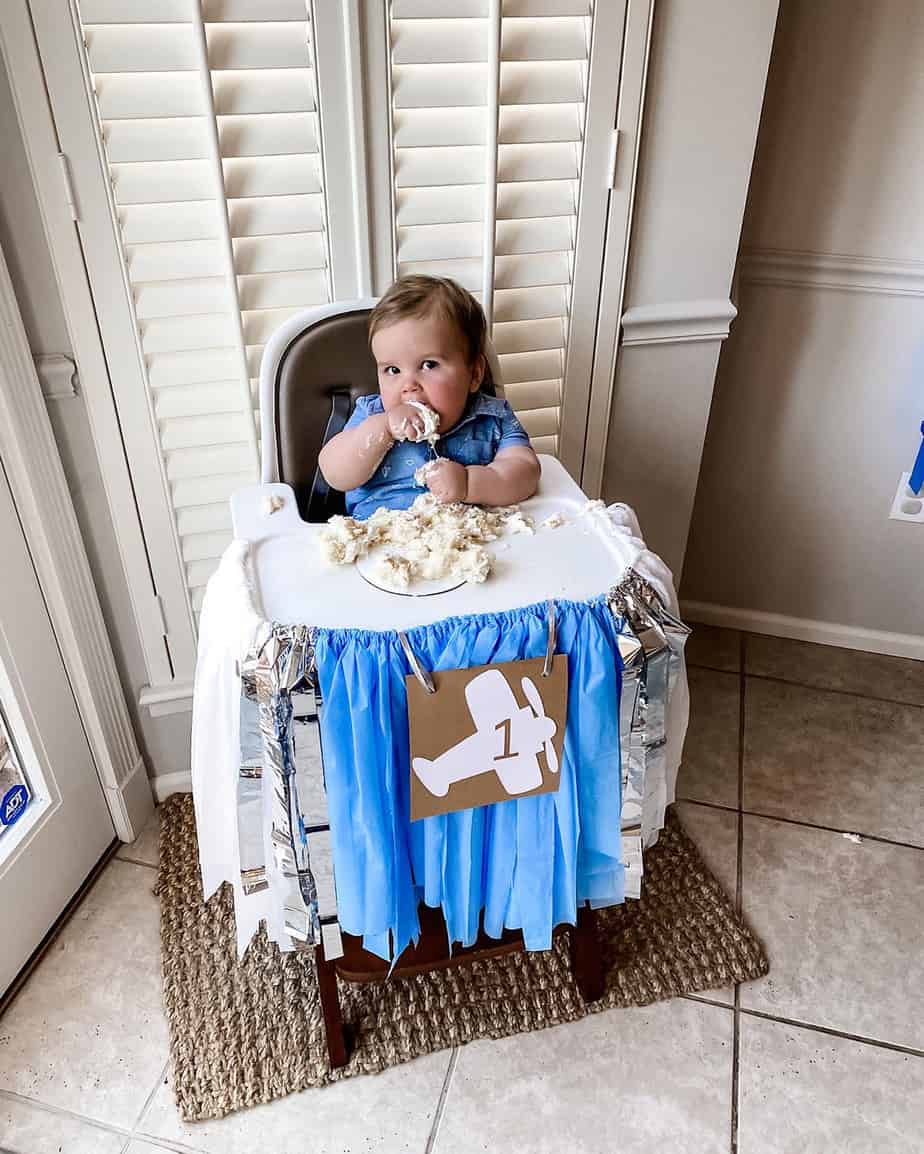 High chair set for first birthday party with blue and white banner hanging and picture of airplane on the front boy eating cake