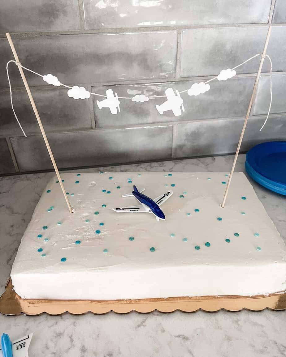 White sheet cake covered in blue sprinkles with blue toy plane. Paper plane and cloud banner made with white paper and hanging from tall skewers.