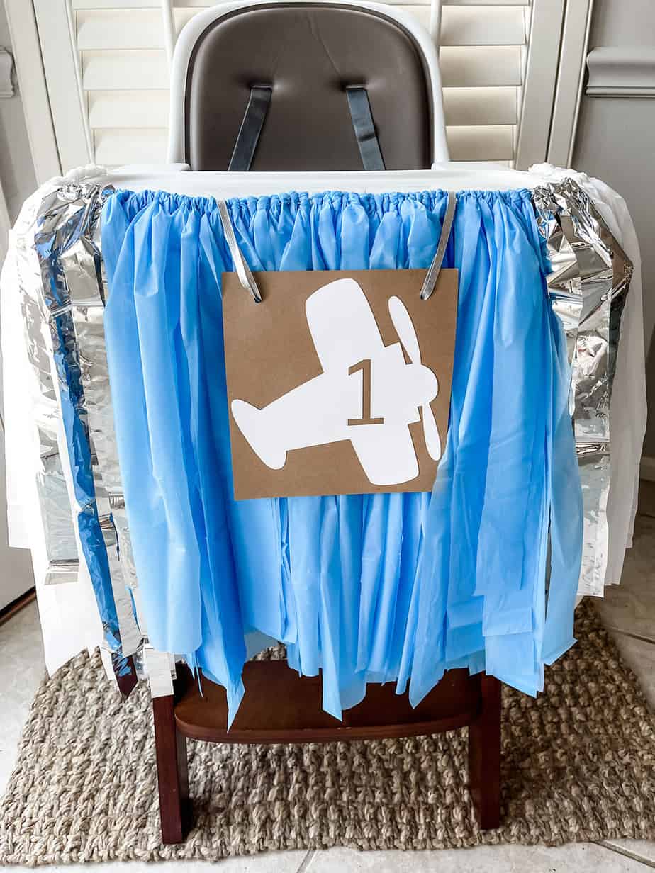 High chair set for first birthday party with blue and white banner hanging and picture of airplane on the front