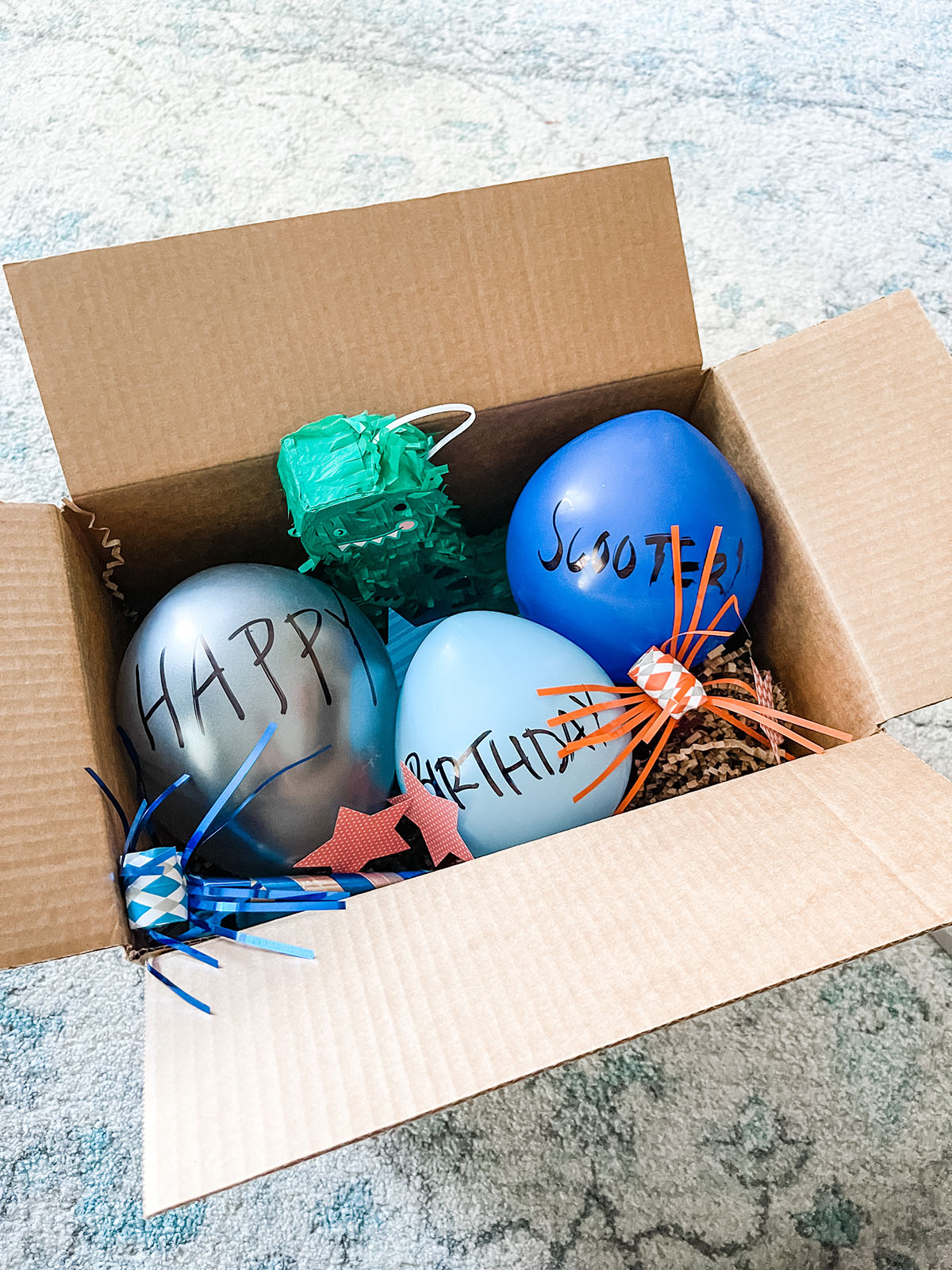 Brown box filled with birthday decorations, dinosaur pinata, blue balloons, and party blowers sitting on a blue and white rug.