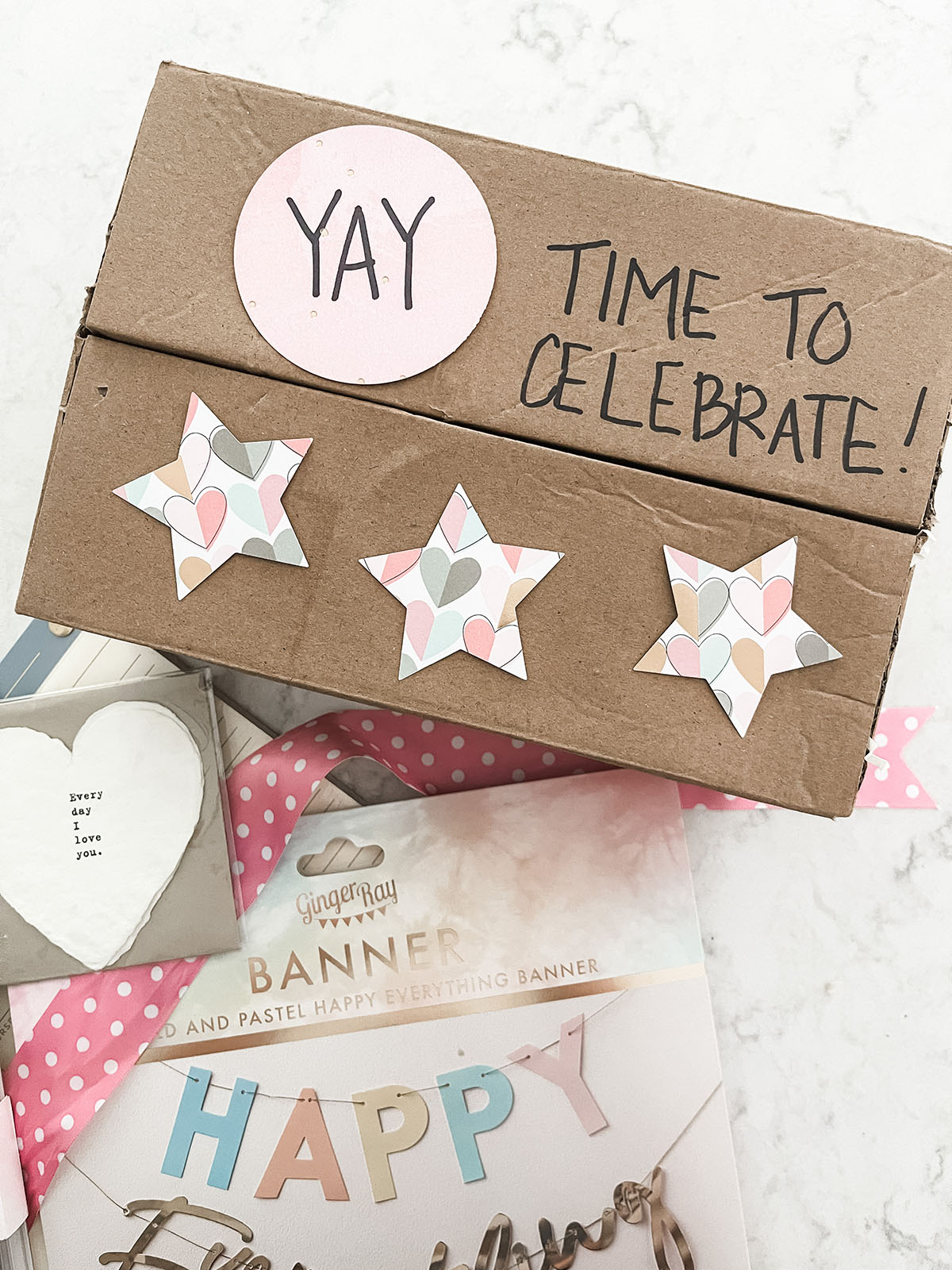 Brown box decorated with paper cut outs in the shape of stars laying next to pink polka dot ribbon and happy birthday banner sitting on white kitchen island.
