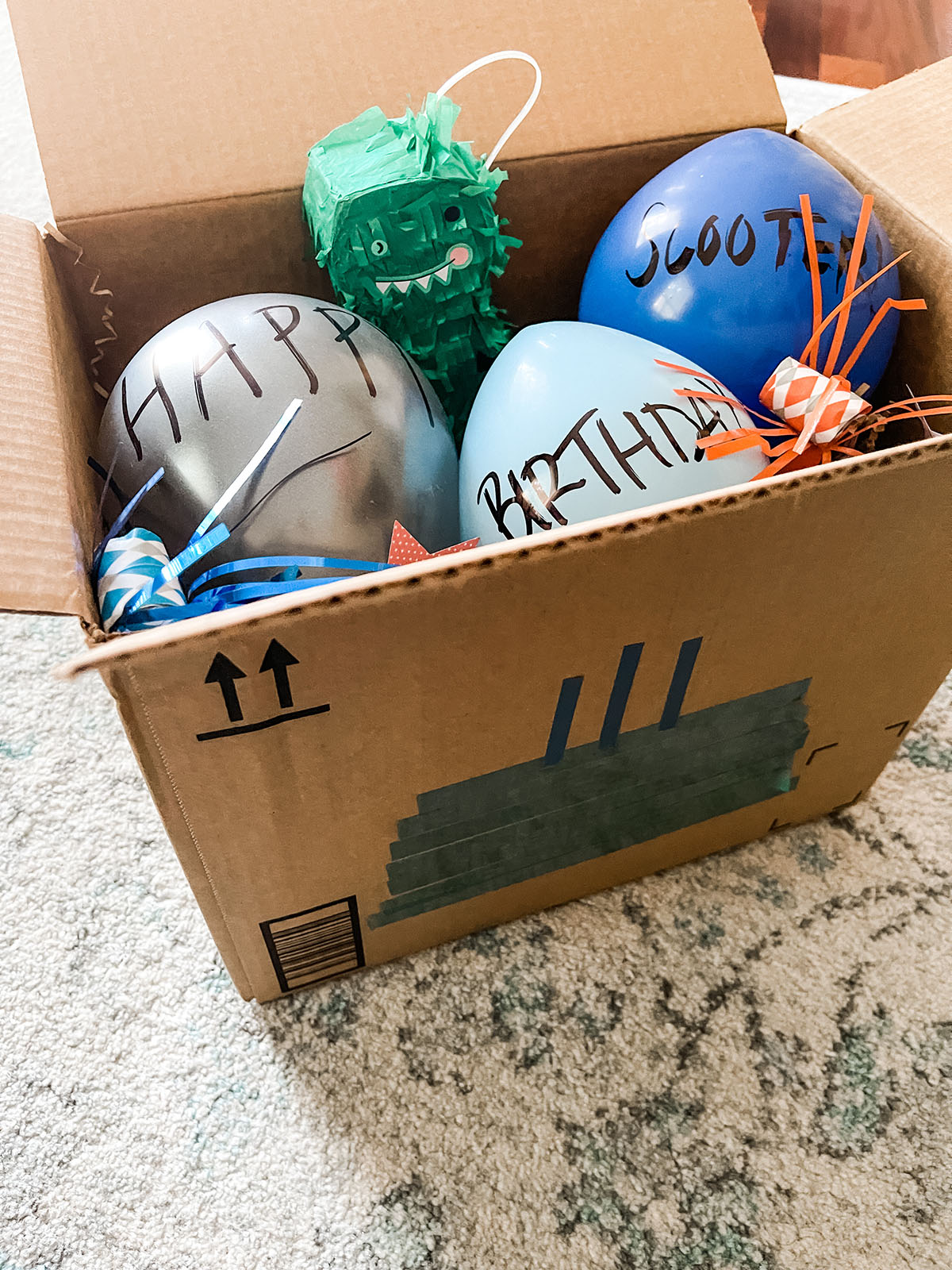 Brown box filled with birthday decorations, dinosaur pinata, blue balloons, and party blowers sitting on a blue and white rug.