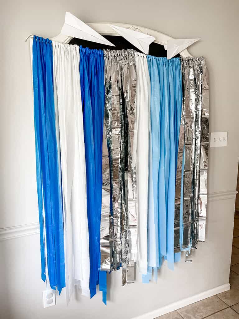 DIY airplane themed party backdrop made from white, blue, and silver paper tablecloth strips.