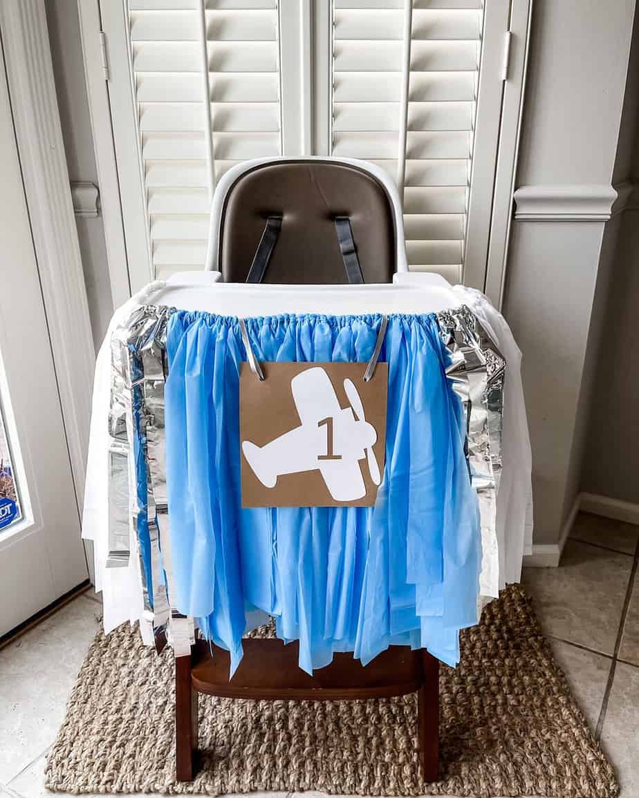 High chair with blue, silver and white banner and #1 airplane sign on front sitting on jute rug ready for a first birthday party.