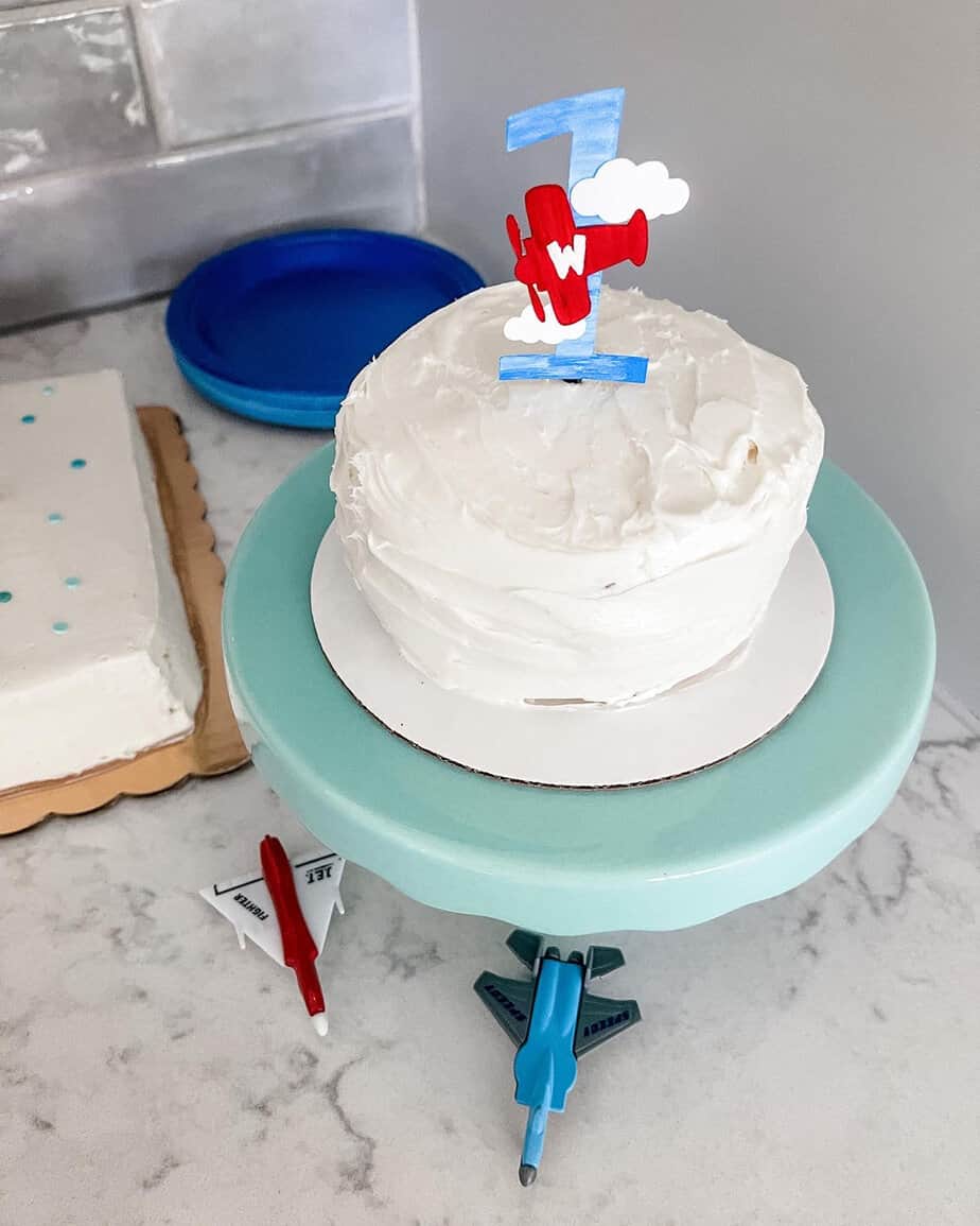White smash cake on a blue cake tier with #1 cake topper that is blue with white clouds and a red airplane on it.