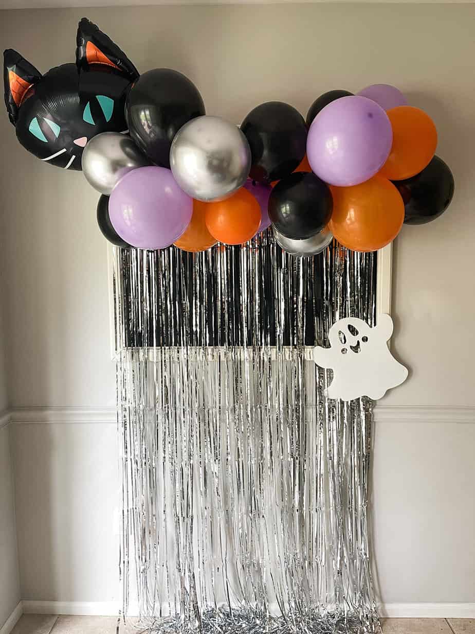 Orange, purple and black halloween balloons on a diy garland with black cat balloon and white ghost over a silver metallic foil backdrop.