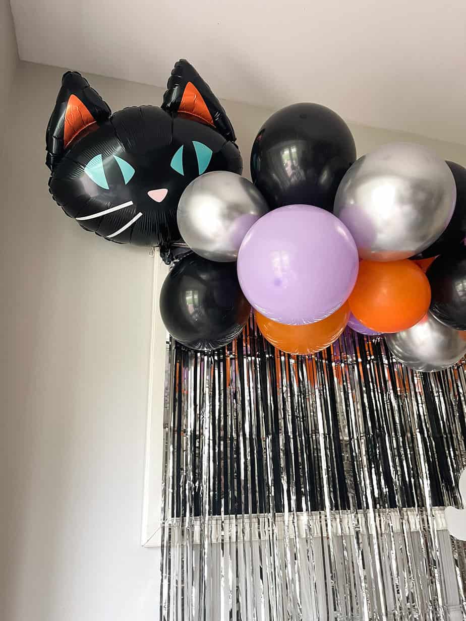 Orange, purple and black halloween balloons on a diy garland with black cat balloon and white ghost over a silver metallic foil backdrop.