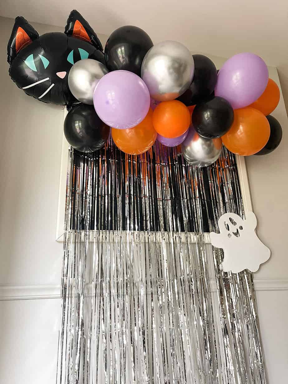 Orange, purple and black halloween balloons on a diy garland with black cat balloon and white ghost over a silver metallic foil backdrop.