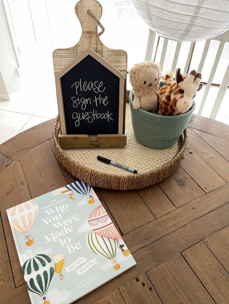 children's book with marker to be signed as a guestbook at a baby shower laying on a wood table with chalkboard sign and green basket holding stuffed giraffe and lion.