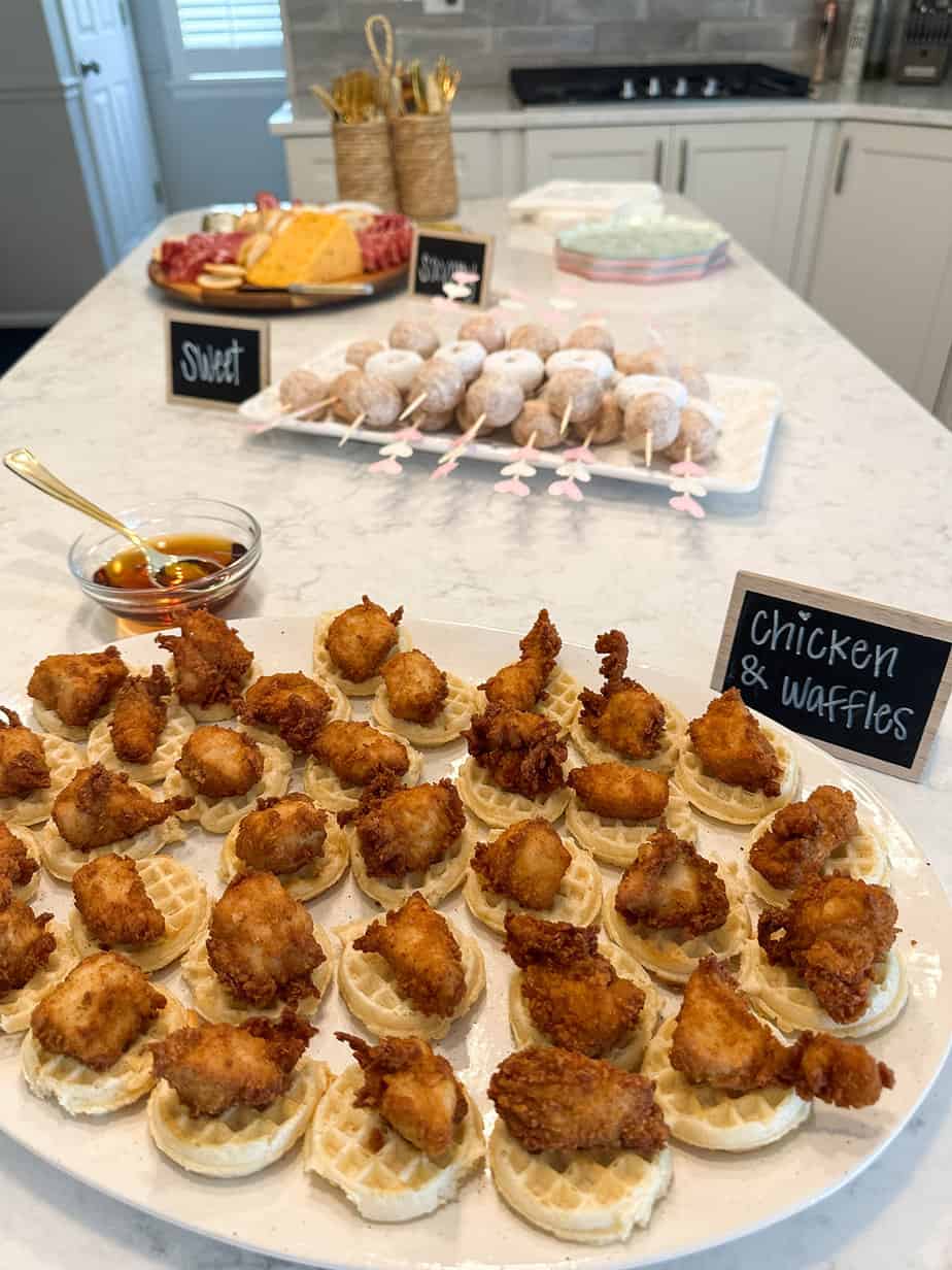 Mini chicken and waffles laying on a large platter with donut skewers and charcuterie in the background set up for a baby shower.