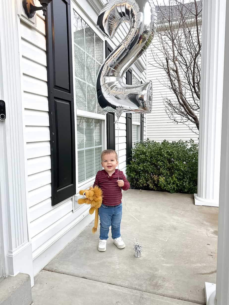 boy holding "2" silver balloon on a front porch dressed in jeans with a stuffed lion