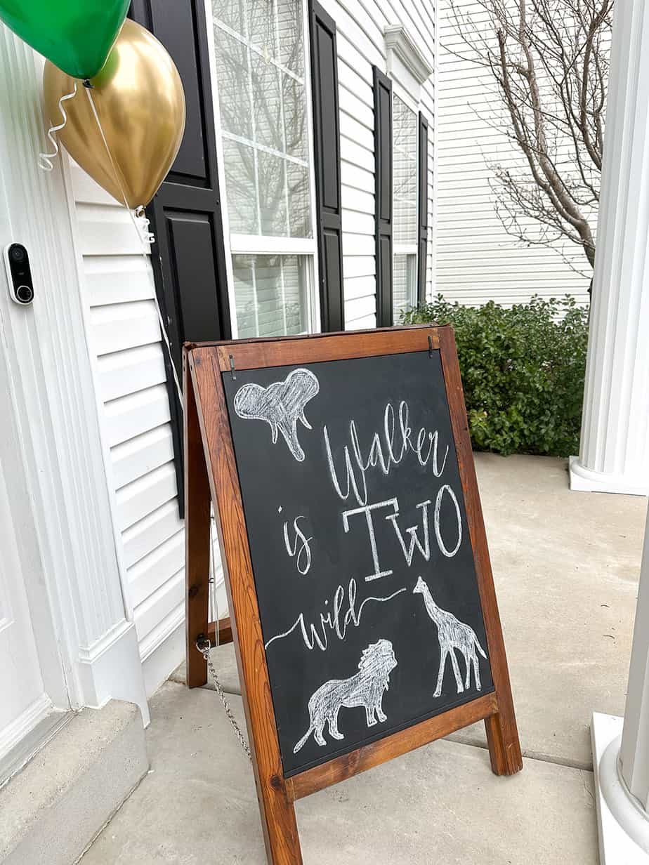 chalkboard sitting on front porch that reads "two wild" with animal silhouettes of an elephant, lion and giraffe drawn on, green and gold balloons attached to the chalkboard.