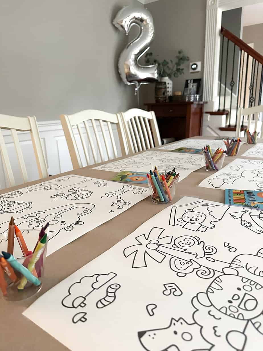 dining table set with butcher paper and animal coloring pages, cups of colorful crayons and coloring books laid out, big silver "2" balloon in background