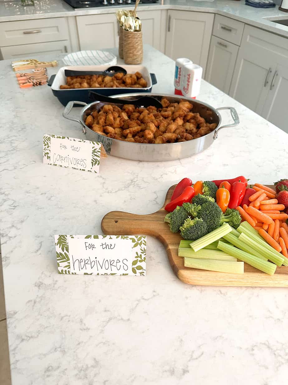 fruit and veggie platter with sign reading "for the herbivores" along with trays of chicken nuggets with sign reading "for the carnivores" all set out on a white kitchen island