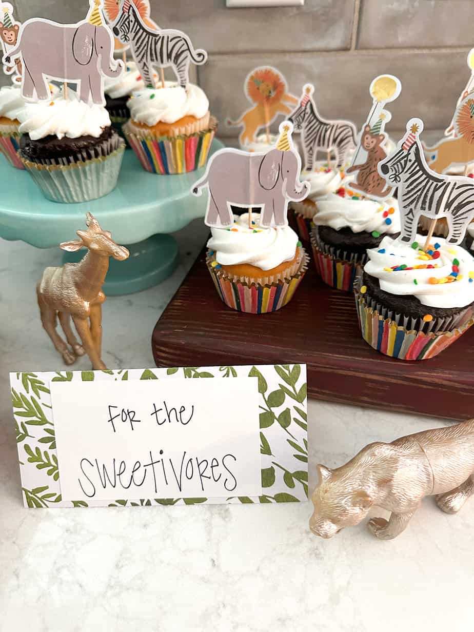 cupcakes with colorful animal toppers wearing birthday hats set on them, sign reading "for the sweetivores" and gold figurine animals of lion and giraffe set nearby for decor